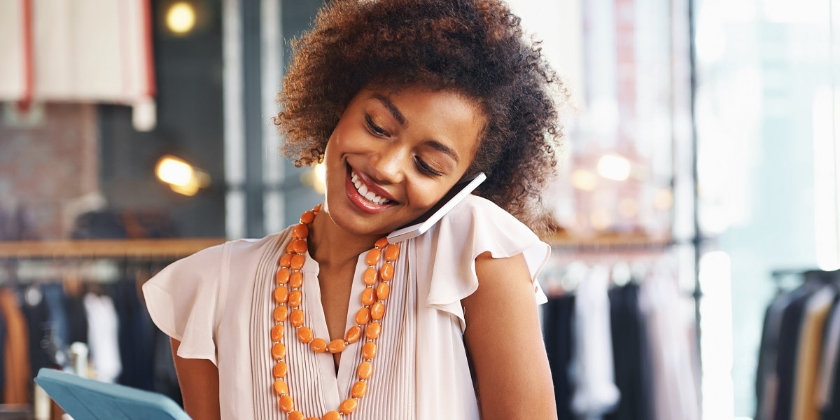 A woman in a blouse with an orange necklace multi-tasking--talking on the phone and on her tablet 