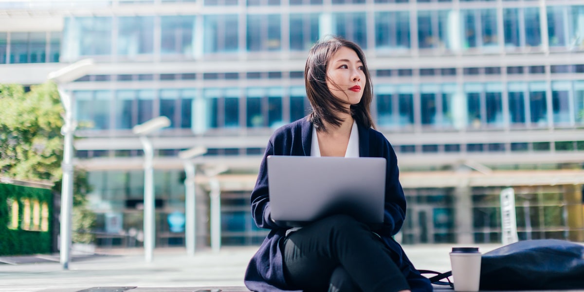 Person sitting outside on a bench with a laptop with an office building in the background.